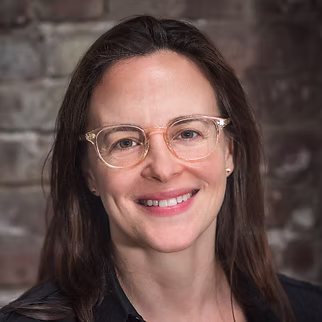 The Freeman Studio A woman with long brown hair and clear glasses smiles at the camera, standing in front of a brick wall outside The Freeman Studio, a renowned New York Acting Studio. New York & Toronto Acting Studio