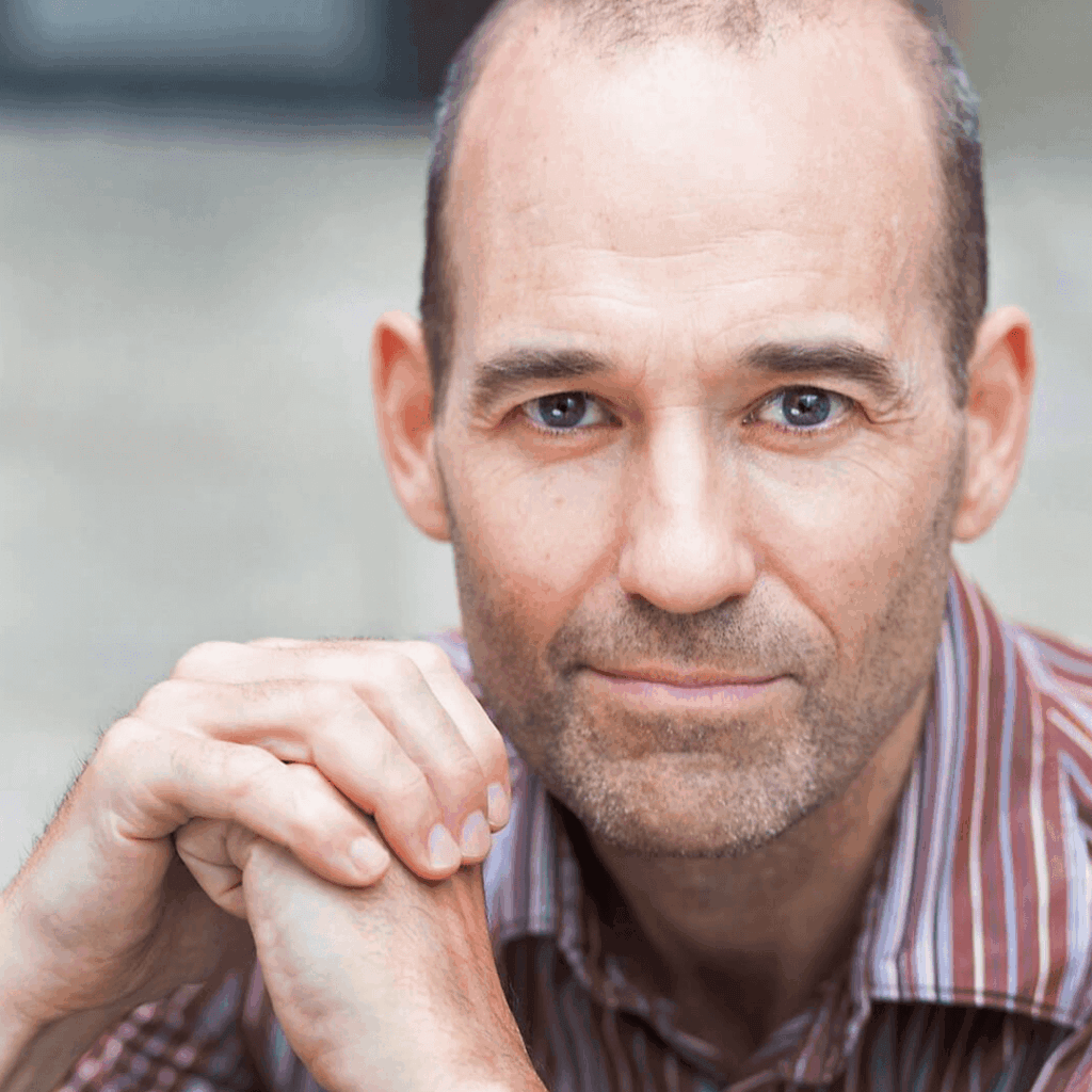 The Freeman Studio A middle-aged man with a receding hairline and stubble rests his hands together, wearing a striped shirt and looking directly at the camera with a neutral expression, evoking the calm focus of a New York Acting Studio student. New York & Toronto Acting Studio