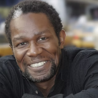 The Freeman Studio A man with short curly hair and a beard smiles at the camera while wearing a black shirt, photographed at The Freeman Studio, with a blurred background behind him. New York & Toronto Acting Studio