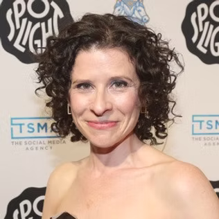 The Freeman Studio A woman with curly dark hair wearing a strapless top smiles at an event with a step-and-repeat backdrop featuring various logos, including the Toronto Acting Studio. New York & Toronto Acting Studio