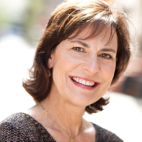 The Freeman Studio Woman with short brown hair smiling outdoors, wearing a dark textured top and a gold necklace, in soft natural light with a blurred background. New York & Toronto Acting Studio