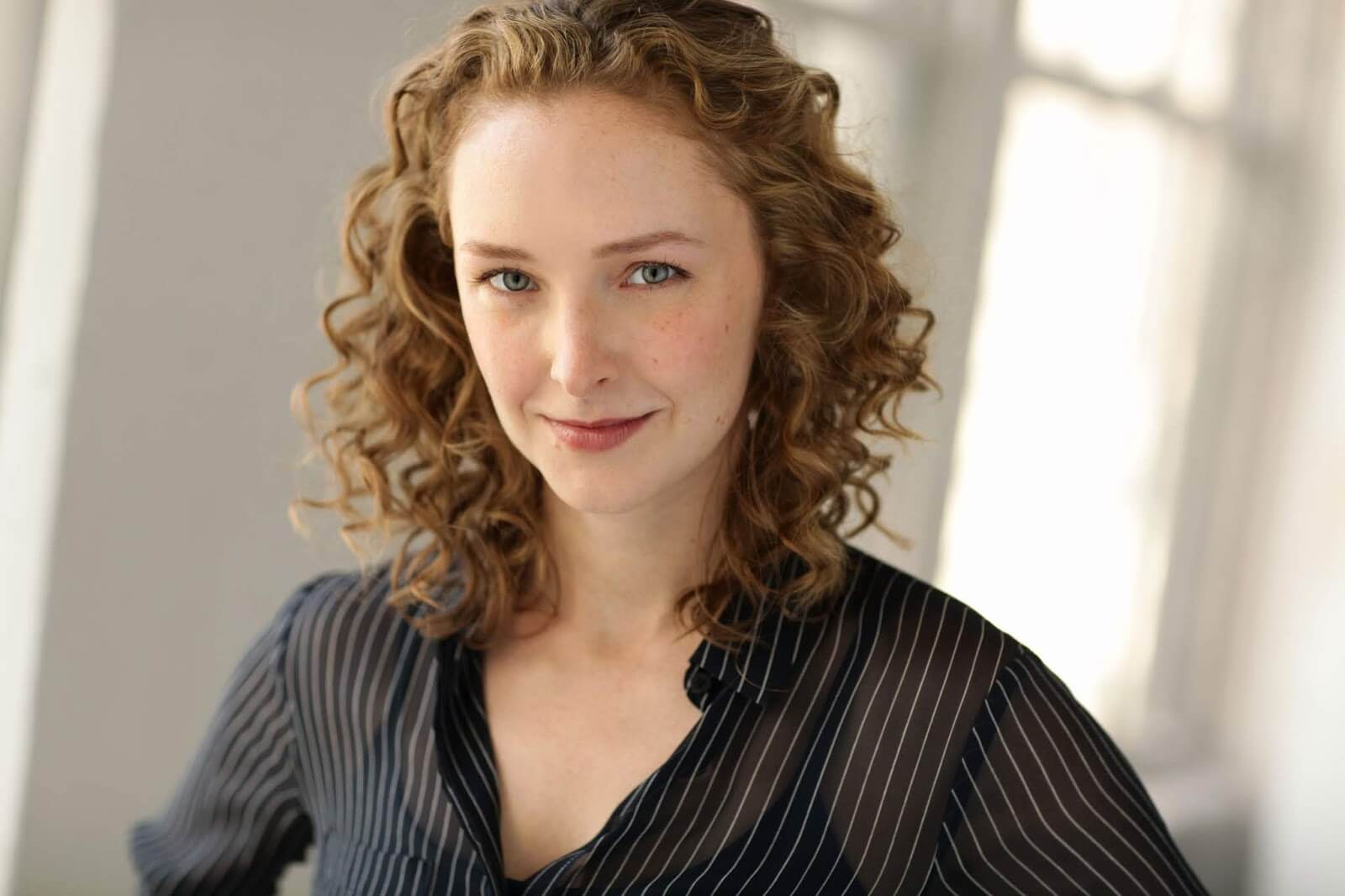 The Freeman Studio A woman with curly light brown hair and fair skin, wearing a black, pinstriped blouse, stands in front of a softly lit, neutral background. New York & Toronto Acting Studio