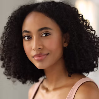 The Freeman Studio A woman with medium-dark skin tone and curly black hair looks at the camera, wearing a light pink top and small hoop earrings, in a softly lit indoor setting at The Freeman Studio. New York & Toronto Acting Studio