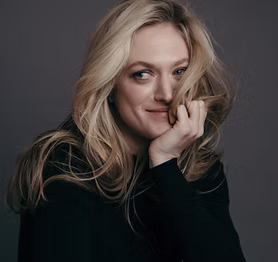 The Freeman Studio A woman with long blonde hair wearing a black top looks at the camera, partially covering her face with her hand, against a plain dark background—a striking portrait from The Freeman Studio. New York & Toronto Acting Studio