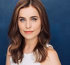 The Freeman Studio A woman with long brown hair and blue eyes wearing a sleeveless white top poses in front of a plain blue background. New York & Toronto Acting Studio