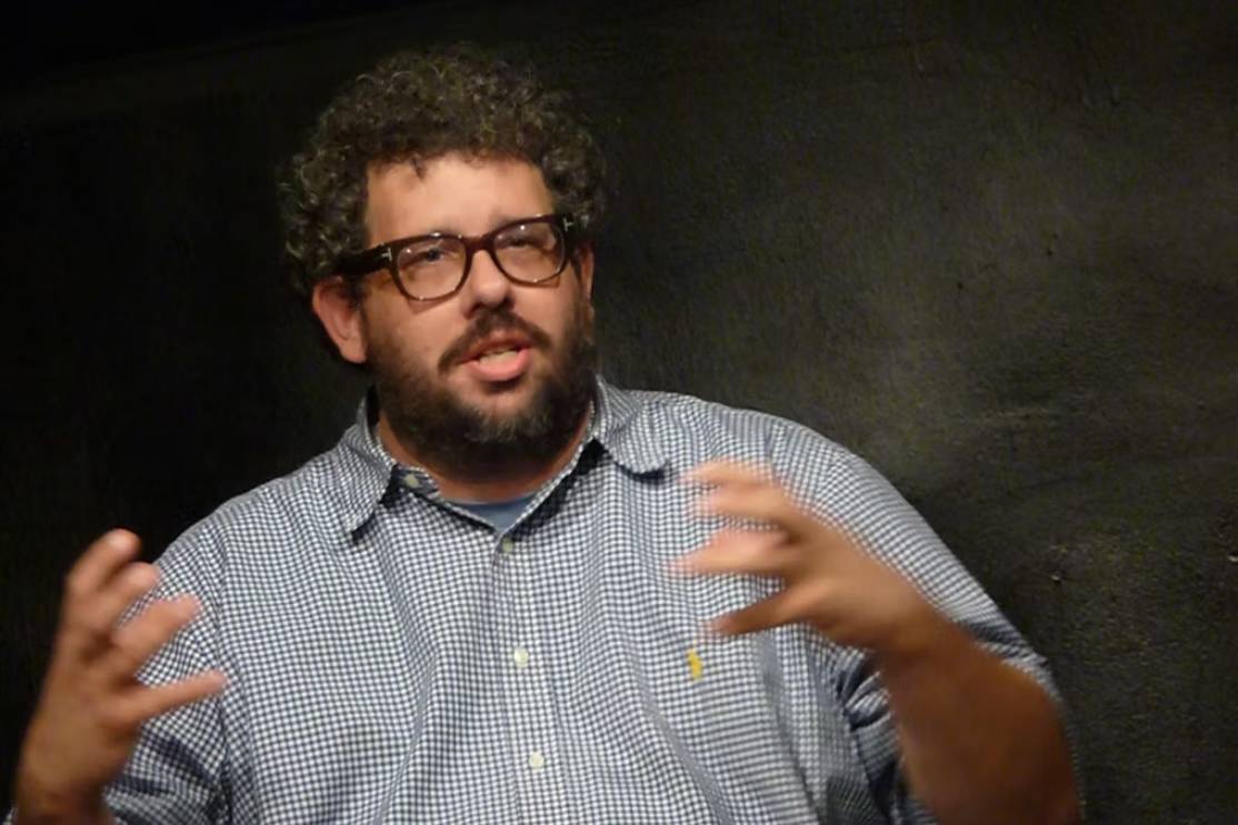 The Freeman Studio A man with curly hair, glasses, and a beard speaks while gesturing with his hands. He is wearing a checkered shirt and is seated against a dark background. New York & Toronto Acting Studio