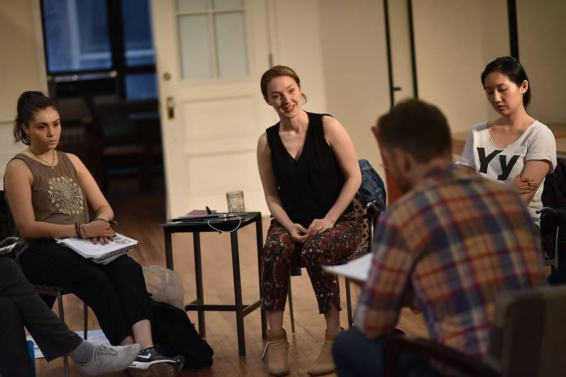 The Freeman Studio Four people sit in a circle engaged in discussion in a well-lit room; papers and notebooks are visible, suggesting a meeting or workshop setting. New York & Toronto Acting Studio