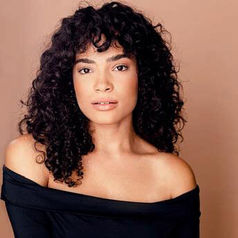 The Freeman Studio Woman with curly dark hair and medium skin tone wearing an off-the-shoulder black top, posing in front of a light brown background. New York & Toronto Acting Studio