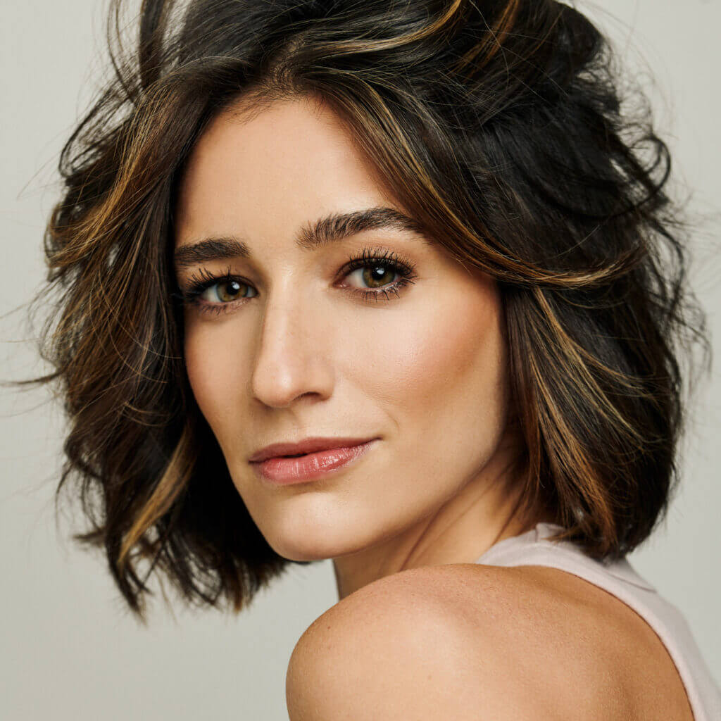The Freeman Studio Woman with short, wavy brown hair with highlights, wearing a sleeveless light top, looking over her shoulder at the camera against a neutral background. New York & Toronto Acting Studio