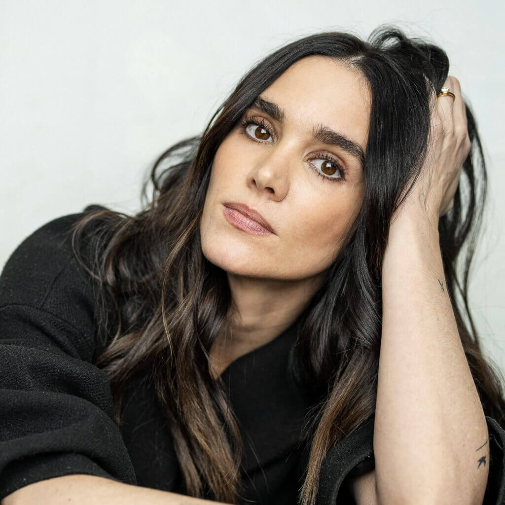 The Freeman Studio A woman with long dark hair and brown eyes rests her head on her hand, looking directly at the camera. She is wearing a black top. New York & Toronto Acting Studio