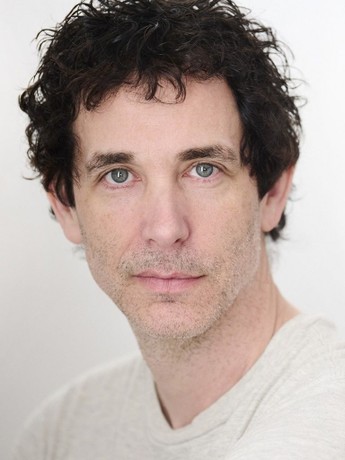 The Freeman Studio A man with short, curly dark hair and light stubble looks directly at the camera. He is wearing a light-colored shirt and the background is plain and light. New York & Toronto Acting Studio