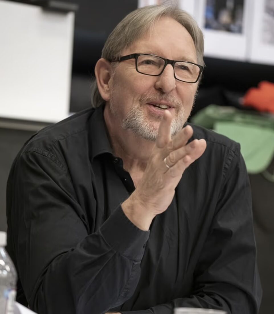 Mark Wing Davey sits at a table with papers, a binder, and a plastic cup, gesturing with his hand while speaking. The Freeman Studio New York & Toronto Acting Studio