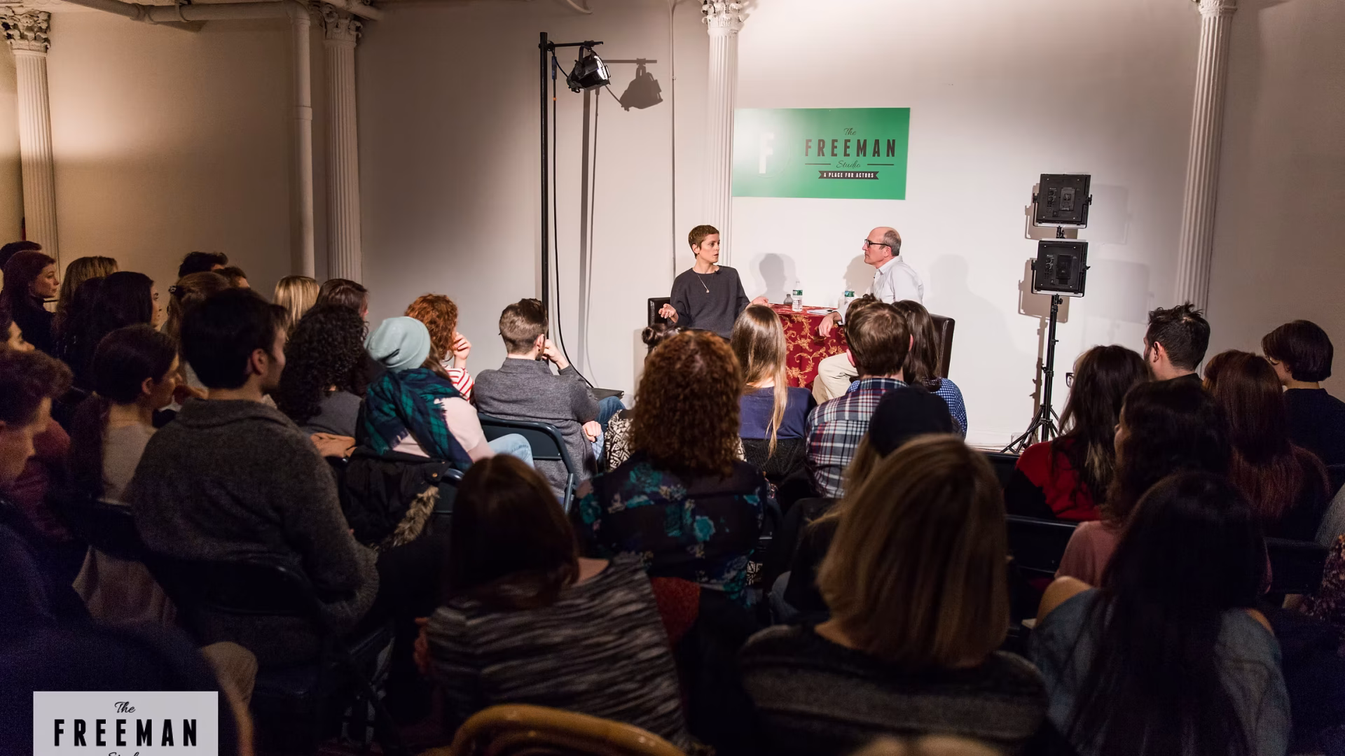 The Freeman Studio Two people sit on a stage in front of an audience during a discussion event at The Freeman Studio, a renowned New York Acting Studio, with a green sign on the wall behind them. New York & Toronto Acting Studio
