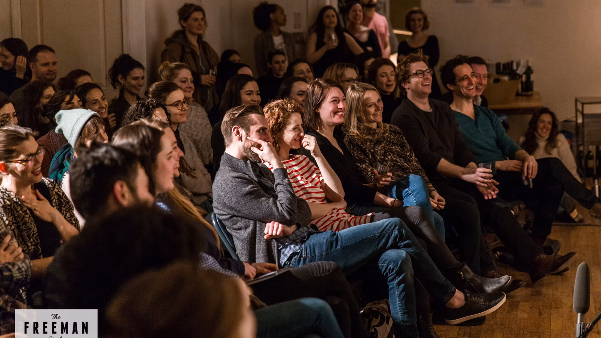 The Freeman Studio A group of people sit closely together in a well-lit room at The Freeman Studio, attentively watching and smiling at an unseen event or performance. New York & Toronto Acting Studio