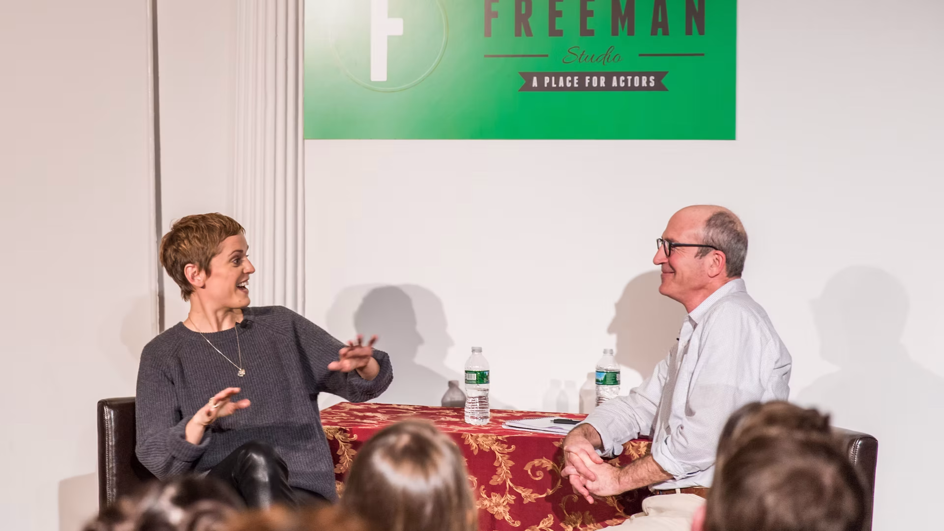 The Freeman Studio Two people sit and converse onstage in front of an audience at The Freeman Studio, a celebrated New York Acting Studio, with a table, two water bottles, and a green sign in the background. New York & Toronto Acting Studio